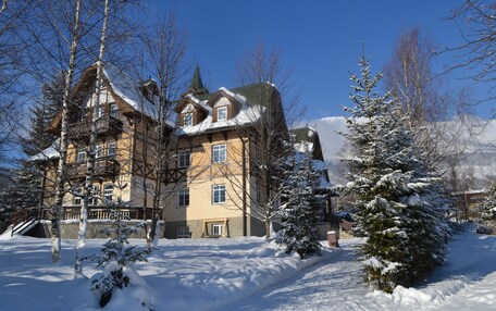 A large, historic building with multiple stories and a snow-covered roof, surrounded by snow-laden trees and a clear blue sky.