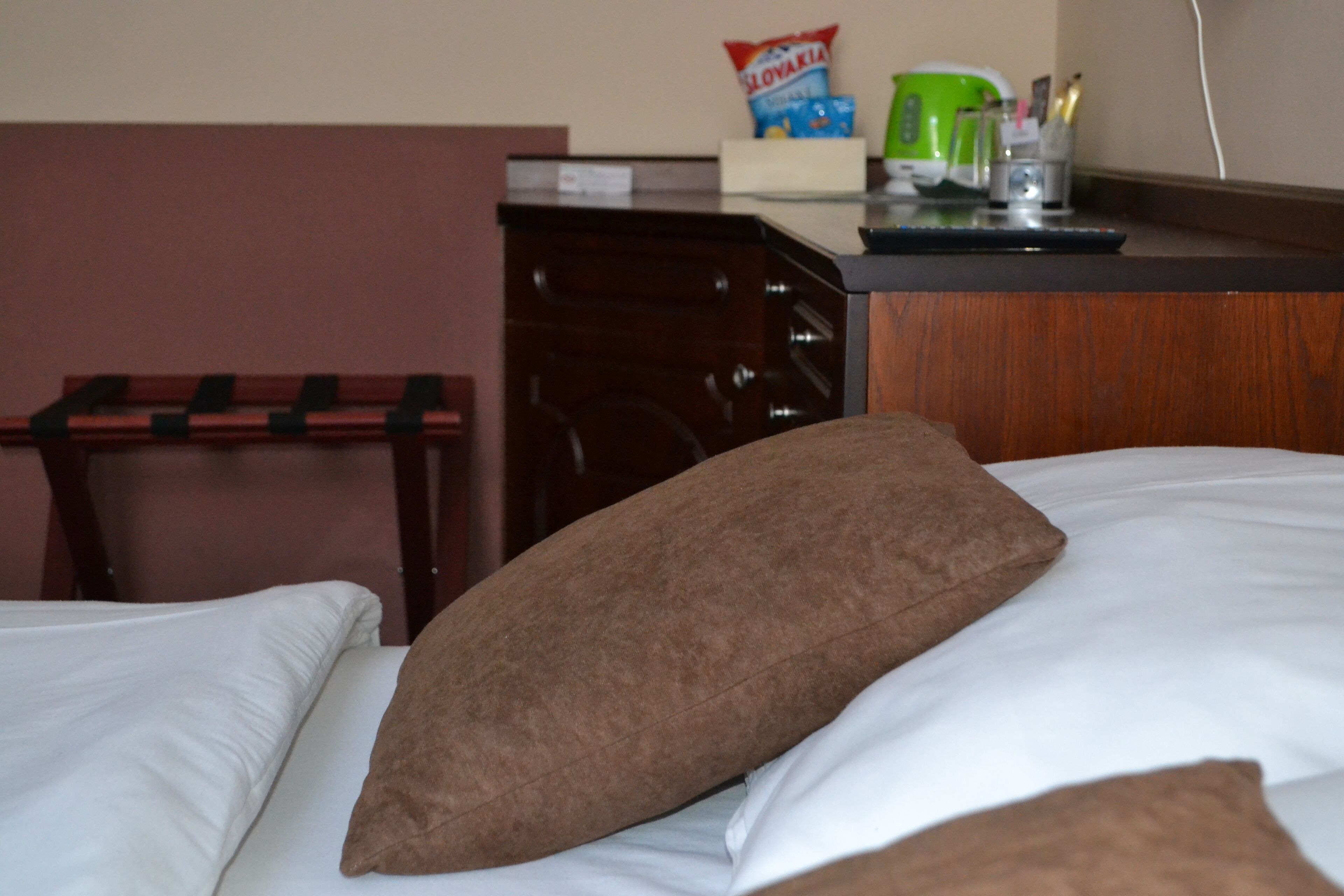 Een hotelkamer met een bed, een bruin kussen, een wit kussen, een houten hoofdbord, een bureau met een groene mok en een zakje snacks.