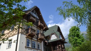 A traditional chalet-style building with multiple balconies and a steeply pitched roof, surrounded by lush greenery and mountains in the background.