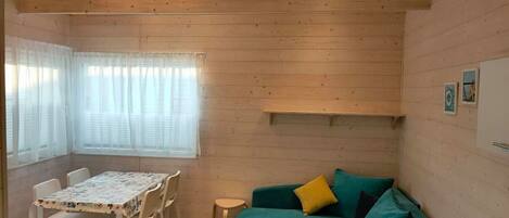 A cozy room with a wooden ceiling, a dining table with chairs, a teal sofa with cushions, and a small round table.