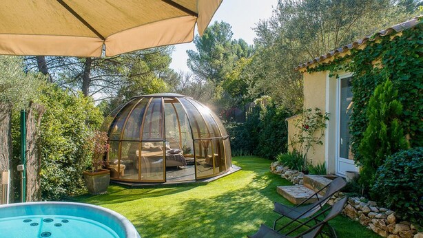 A glass-enclosed hot tub with a patio chair and umbrella, surrounded by a well-manicured lawn and a stone wall. - Domaine Jobert (Aubagne)