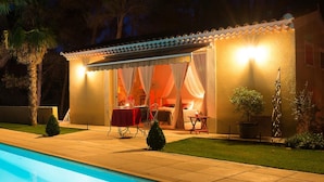 A poolside area with a covered seating area, a pool, and a well-lit building with curtains. - Domaine Jobert (Aubagne)