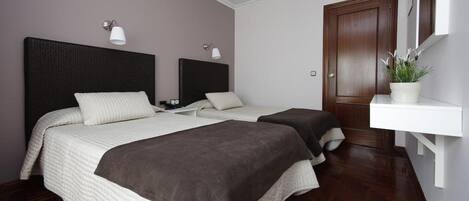 Un dormitorio con dos camas, un cabecero oscuro, un estante blanco con una planta y una puerta de madera.