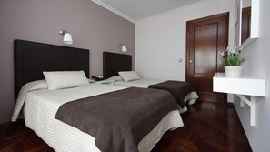 Un dormitorio con dos camas, un cabecero oscuro, un estante blanco con una planta y una puerta de madera.
