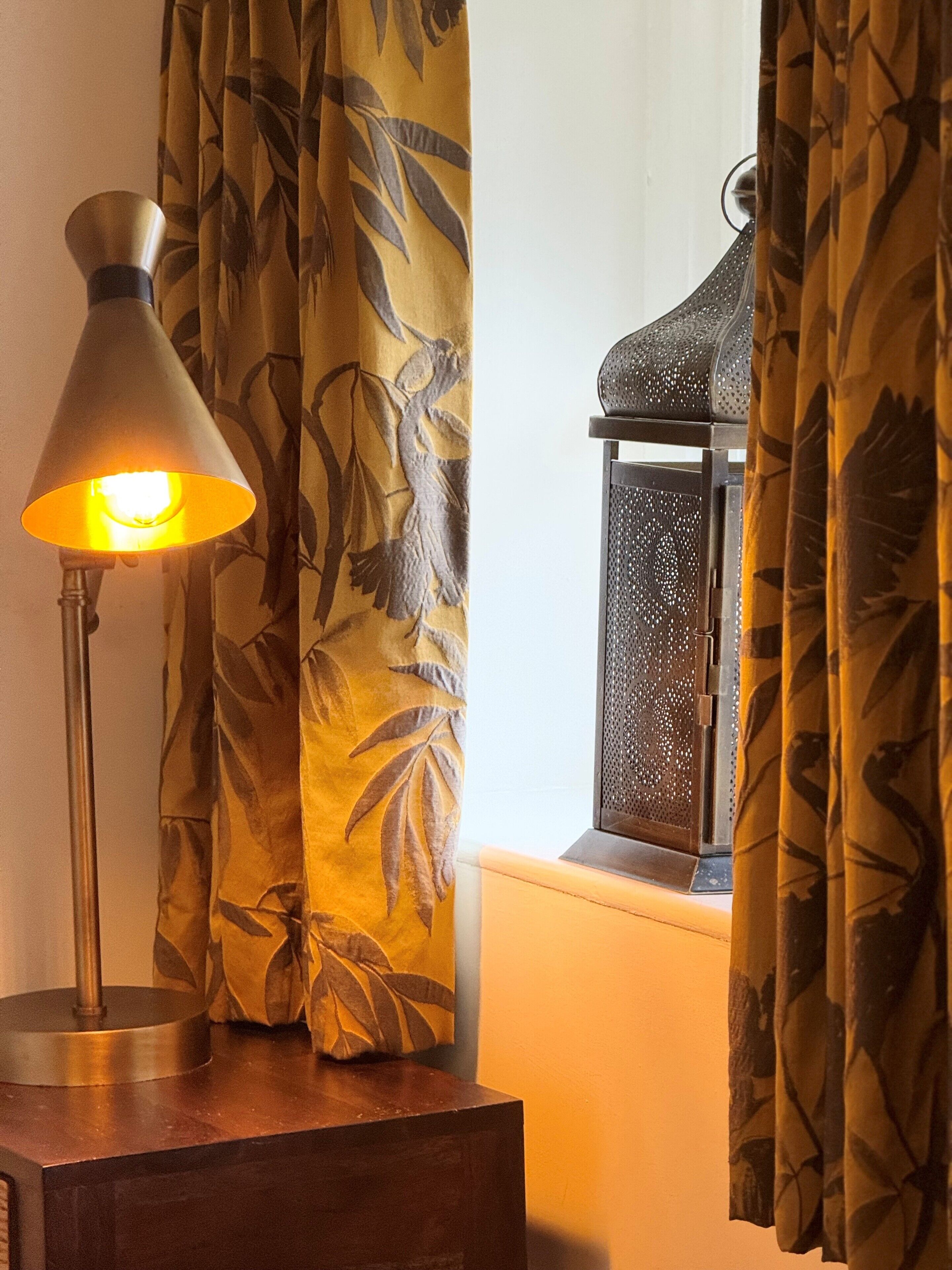 Een lamp op een nachtkastje, een raam met uitzicht en een gordijn met dessin.