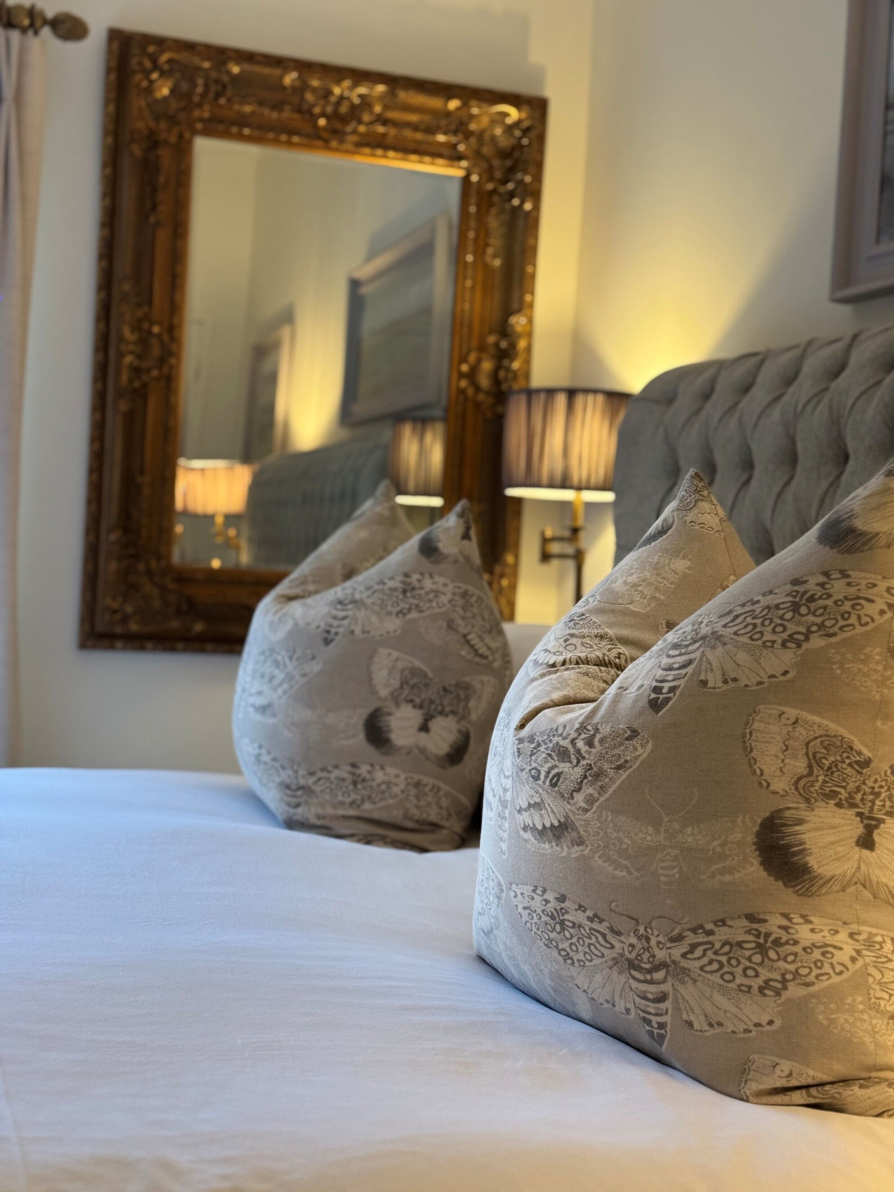 Una cama bien hecha con almohadones estampados, un espejo grande y ornamentado, y una lámpara en una mesita de noche.