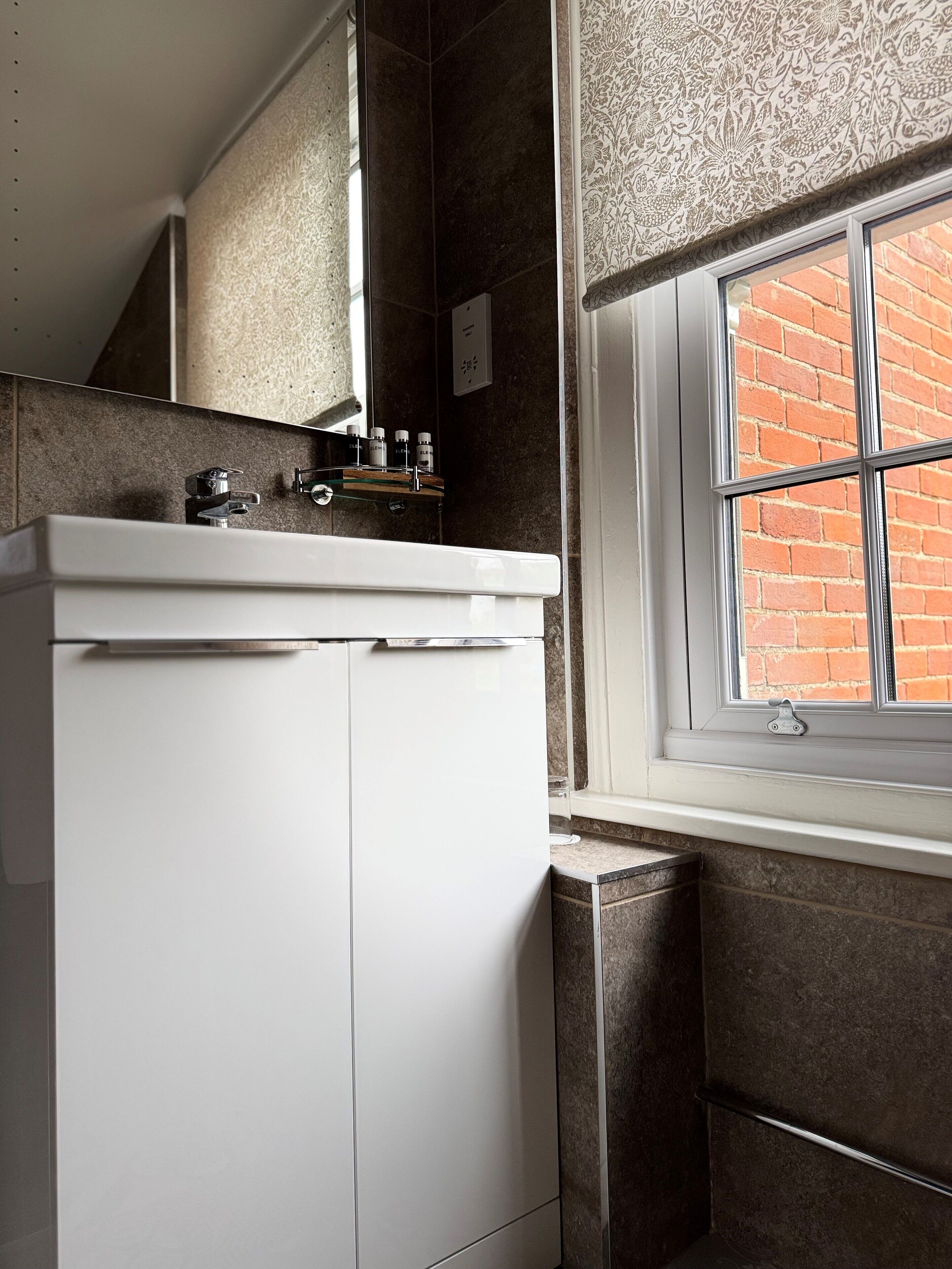 Un baño con un lavabo blanco, un espejo y una ventana que da a un edificio de ladrillo.