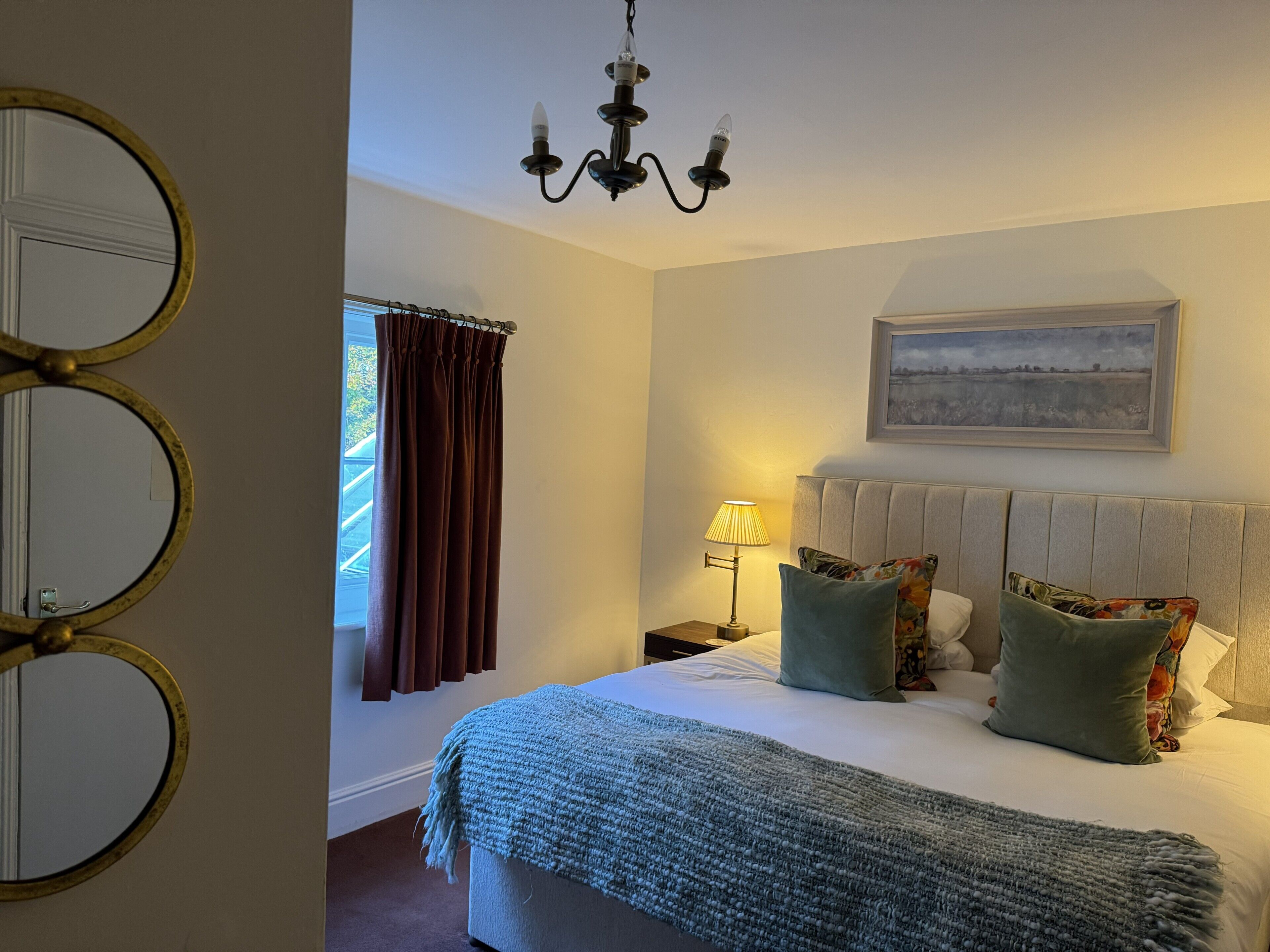 A bedroom with a bed, a bedside lamp, a painting on the wall, and a window with curtains.