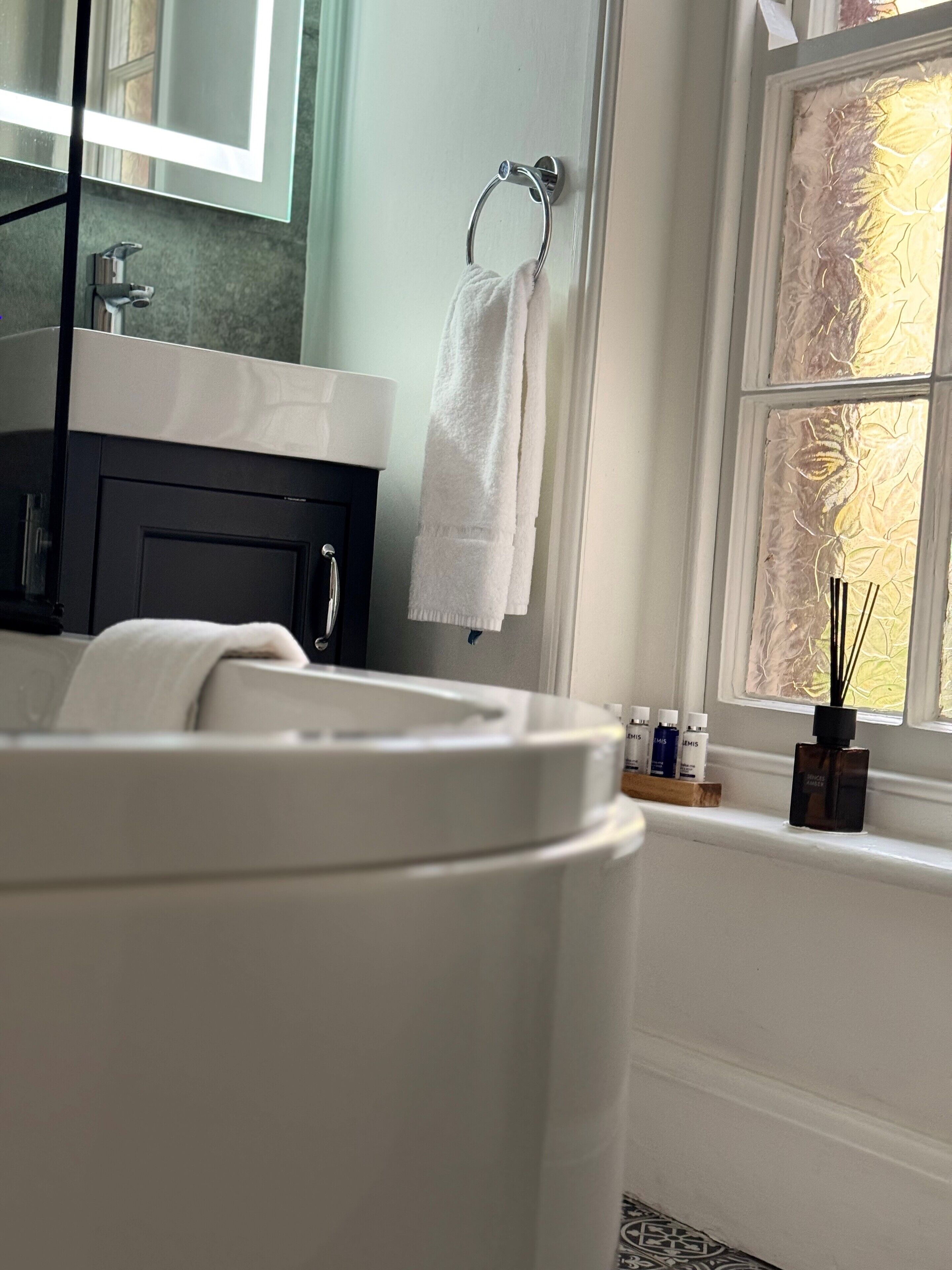 Un baño con una bañera blanca, un tocador negro, un perchero con una toalla blanca y un ventanal con un diseño decorativo.