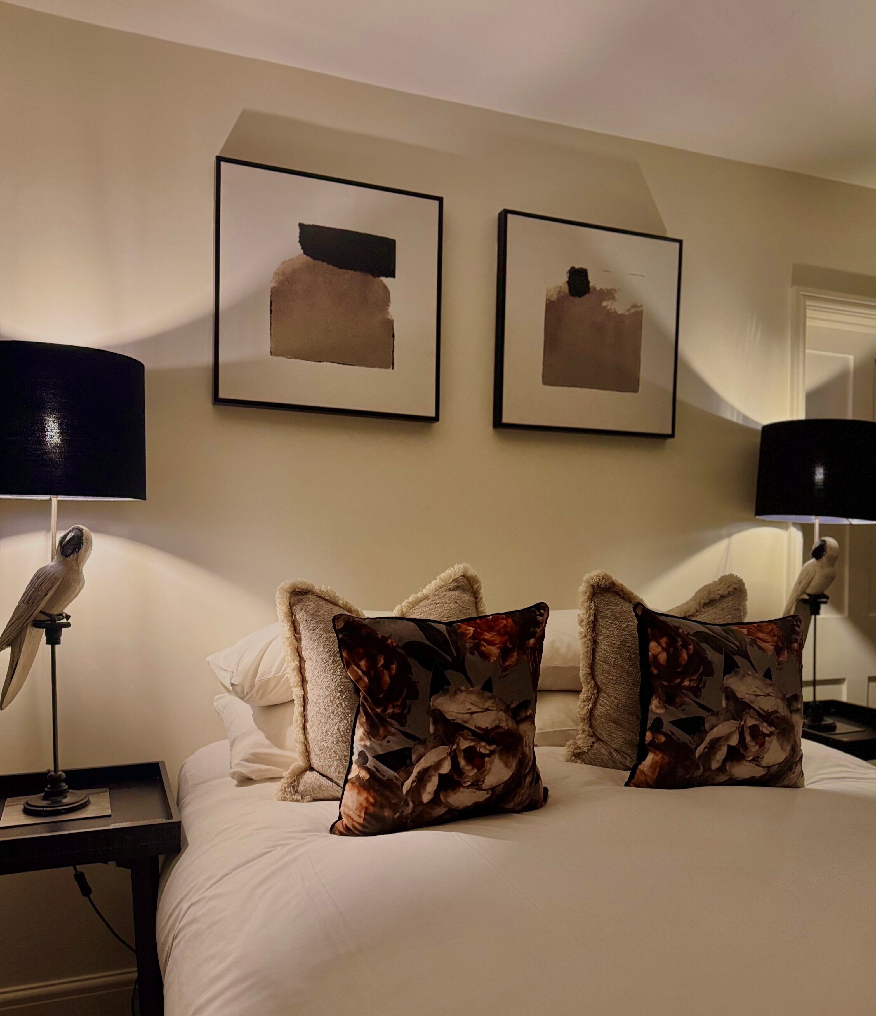 A hotel room with a bed, two pillows, a nightstand, a lamp, and two framed artworks on the wall.