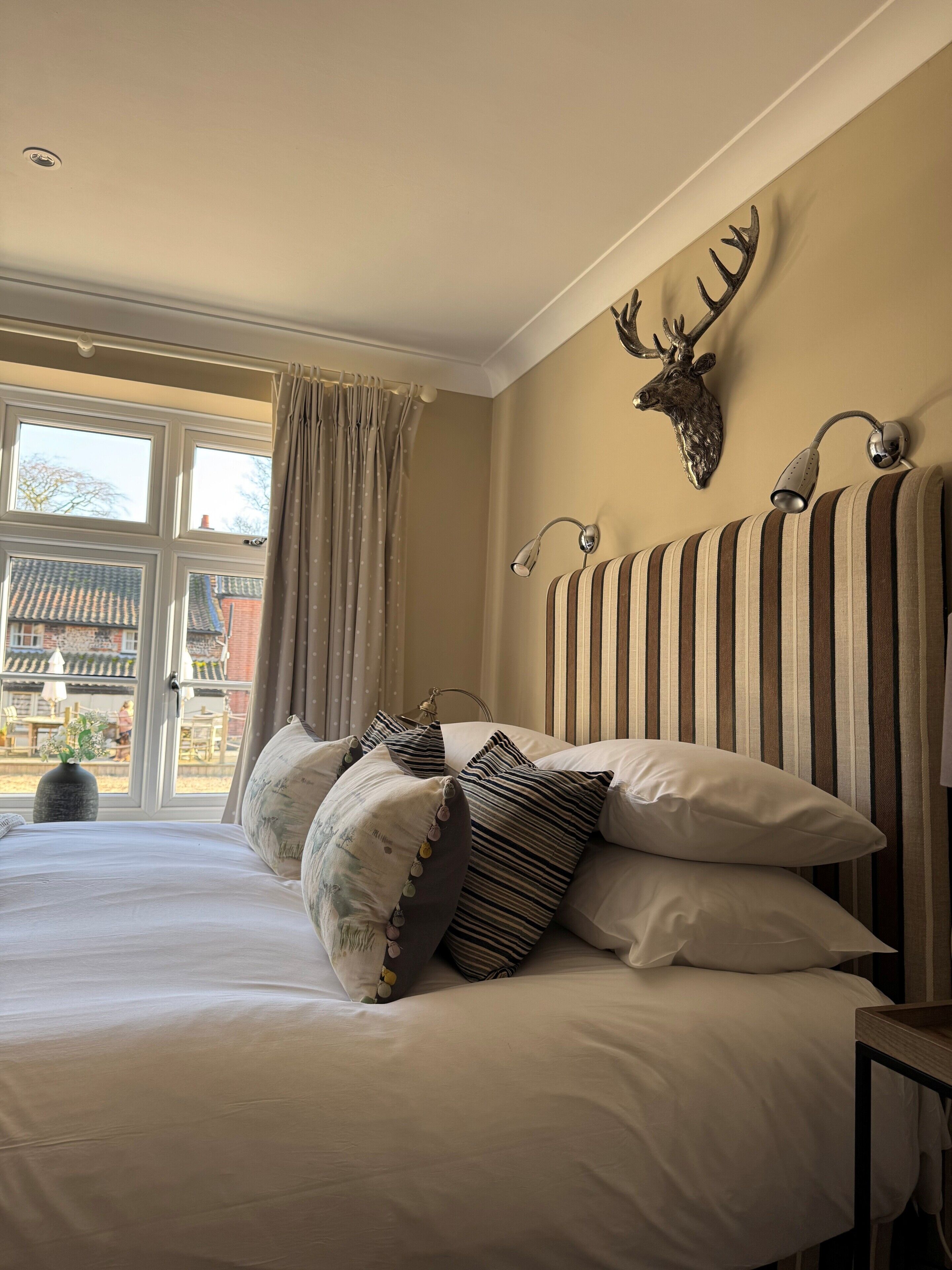 A bed with striped headboard, a mounted deer head, and a window with curtains.