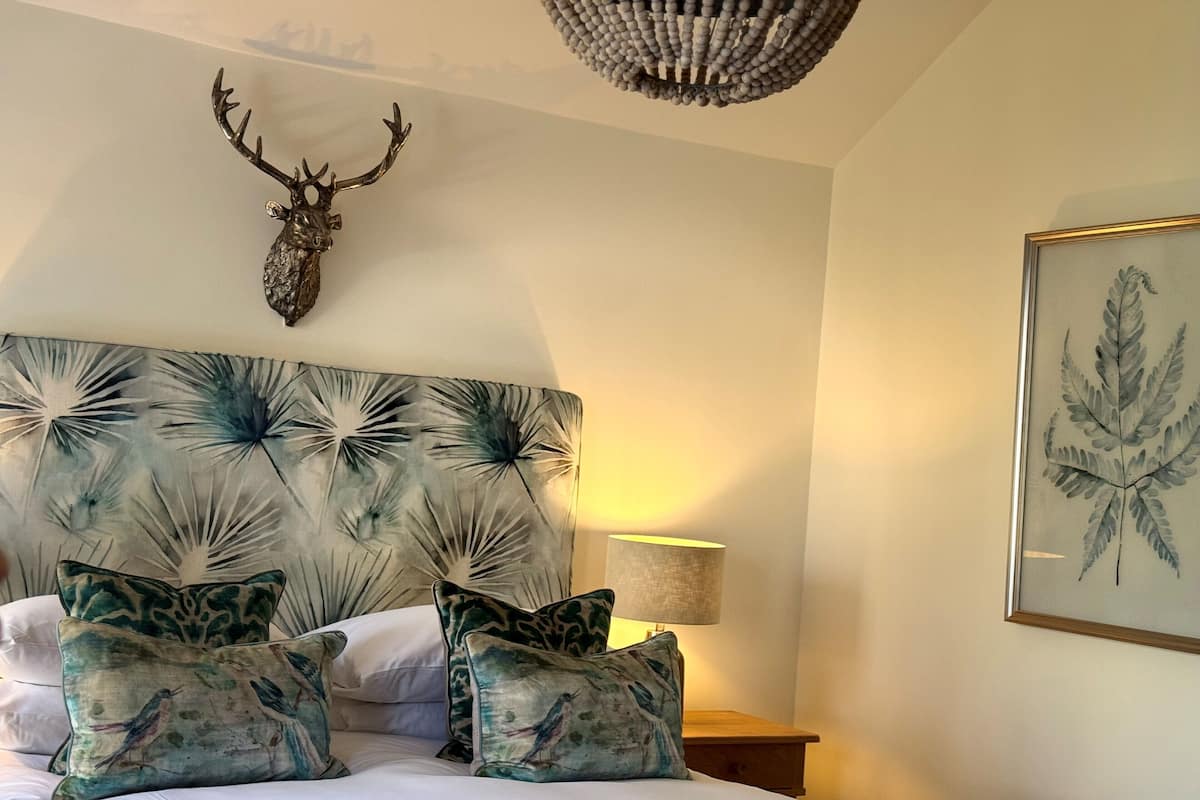 A bedroom with a bed, decorative pillows, a wall-mounted deer head, a framed fern, and a hanging light fixture.