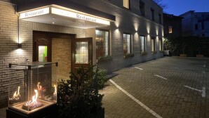 Un edificio con un letrero que dice “Restaurant” sobre la entrada, un cerramiento de vidrio con una chimenea escultural y plantas en macetas a lo largo del sendero.