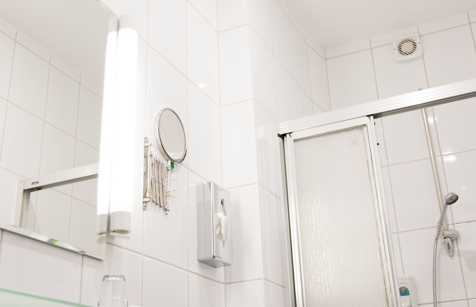 Un baño con azulejos blancos, un espejo, un cabezal de ducha de mano y un dispensador de toallas.
