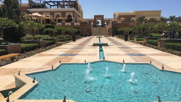Una piscina amplia y cristalina con múltiples fuentes, rodeada por un paseo empedrado y palmeras.