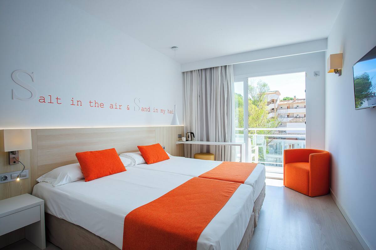 A hotel room with a large bed, two orange pillows, a white headboard, a nightstand, a desk, a chair, a TV, and a balcony with curtains.