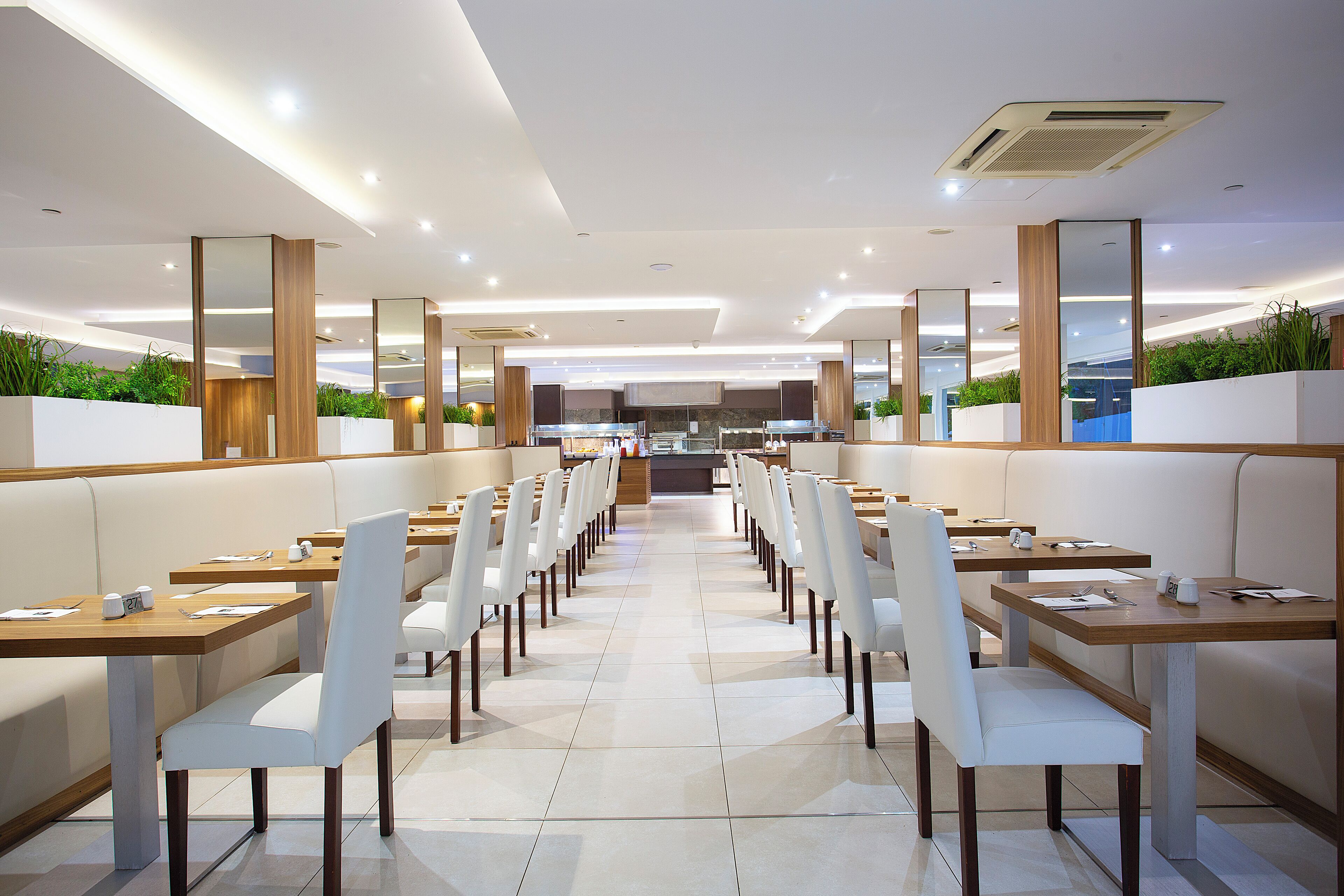 Un restaurant moderne avec de longues tables et des chaises blanches, une cuisine à l’arrière-plan et des plantes sur les murs.