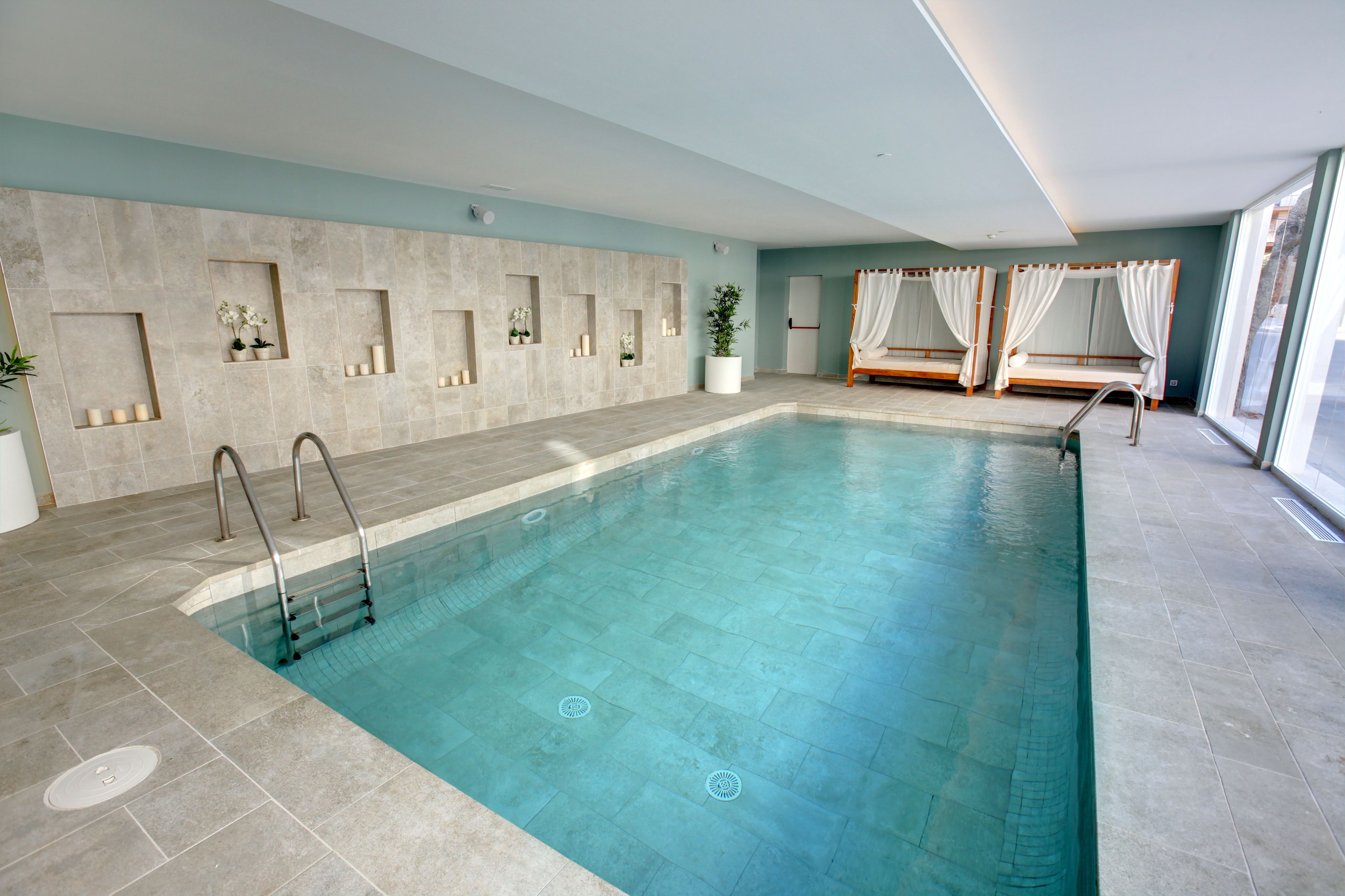 Una gran piscina interior con agua cristalina, paredes de azulejos y una zona de asientos con cortinas blancas.