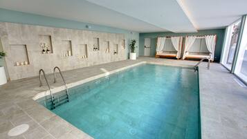 Una gran piscina interior con agua cristalina, paredes de azulejos y una zona de asientos con cortinas blancas.
