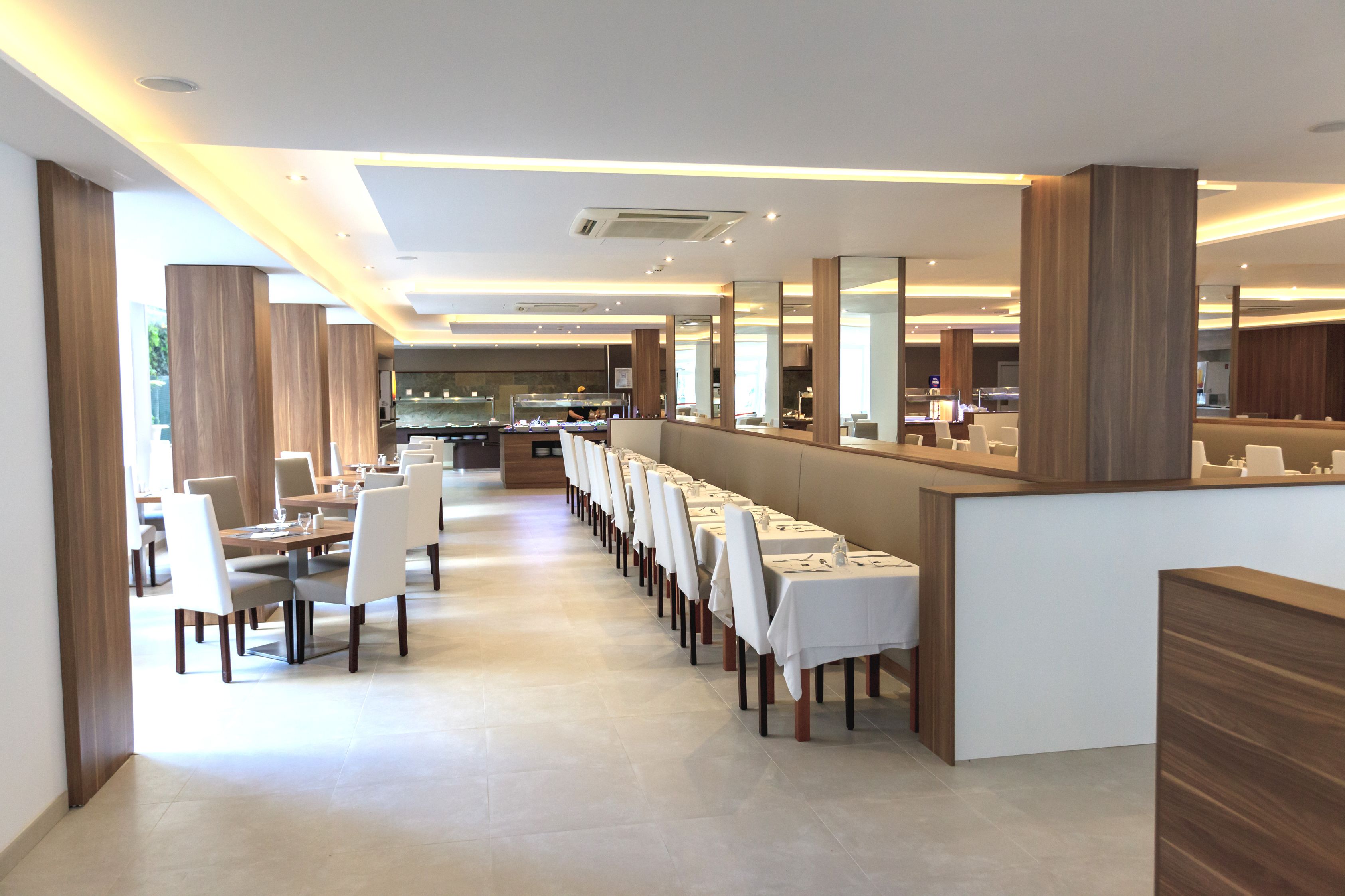 Un'area ristorazione di un hotel moderno con lunghi tavoli, sedie bianche e un bancone centrale.