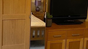 Une chambre avec une armoire en bois, un lit recouvert d’une couette blanche, un téléviseur posé sur un meuble en bois et une petite plante en pot.