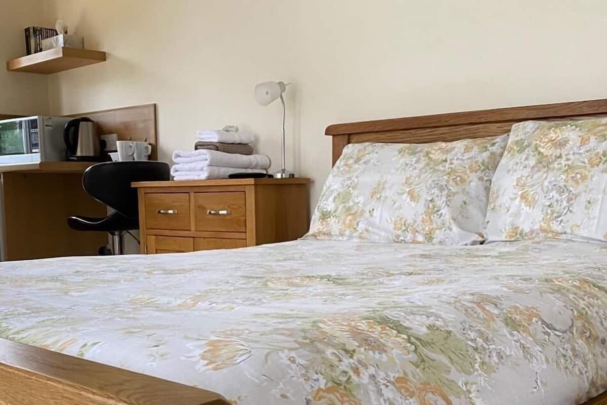 Quarto com estrutura de cama de madeira, roupa de cama floral, mesa de cabeceira com abajur e escrivaninha com chaleira e chávenas.