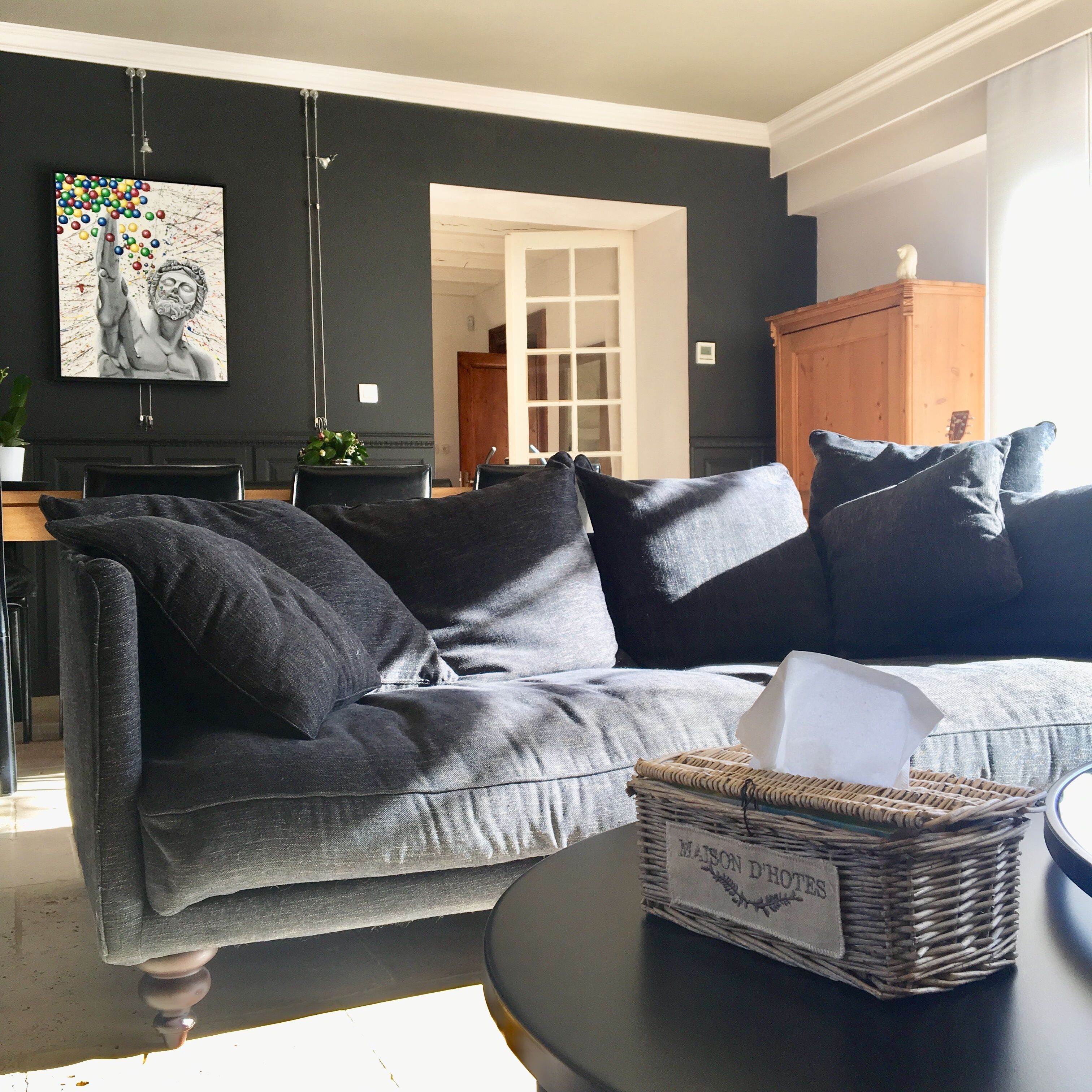 Una sala de estar moderna con un sofá gris oscuro, una cesta de mimbre con la etiqueta "MAISON D'HOTES" y un cuadro en la pared.