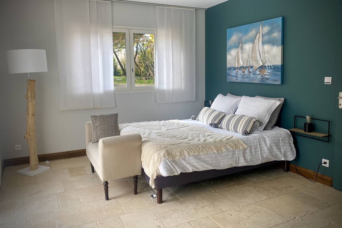 A bedroom with a bed, a chair, a window with curtains, a painting, and a lamp.