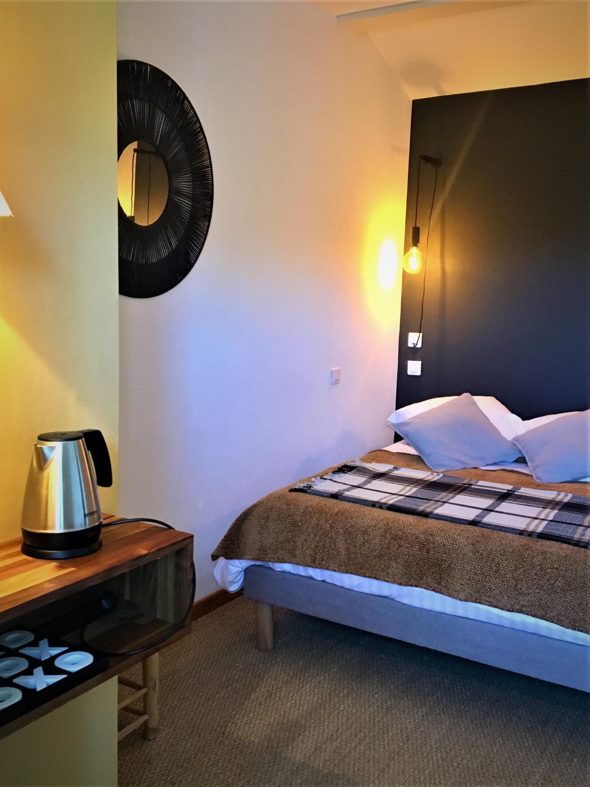 A hotel room with a bed, a bedside table with a kettle, a wall-mounted mirror, and a light fixture.