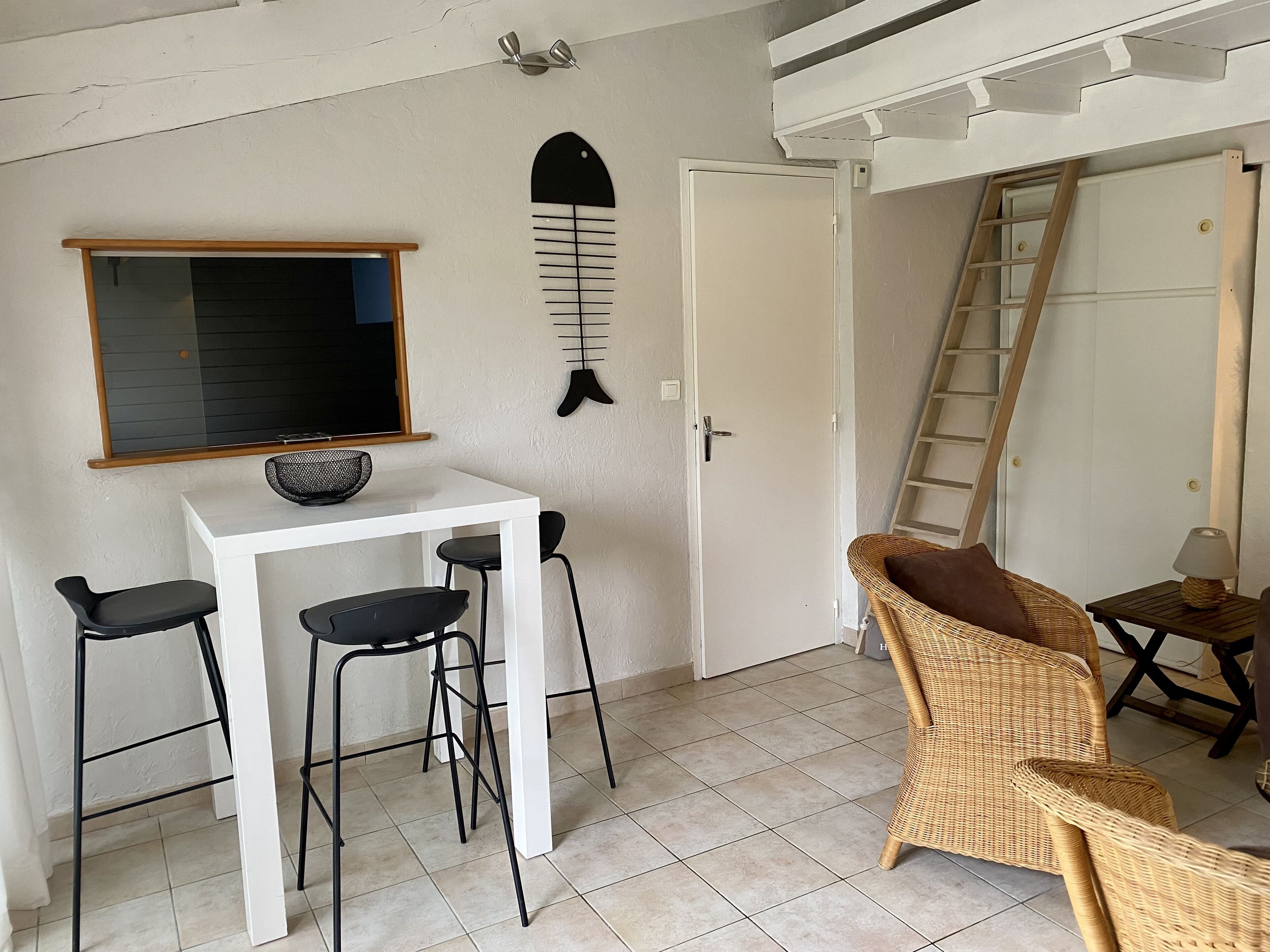 A room with a white table and two black chairs, a wicker chair, a ladder, and a wall-mounted fish decoration.