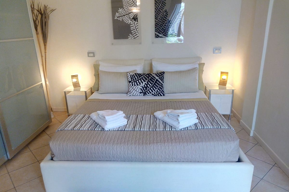 A neatly made bed with a striped bedspread and two pillows, a bedside table with a lamp, and a wall-mounted mirror.