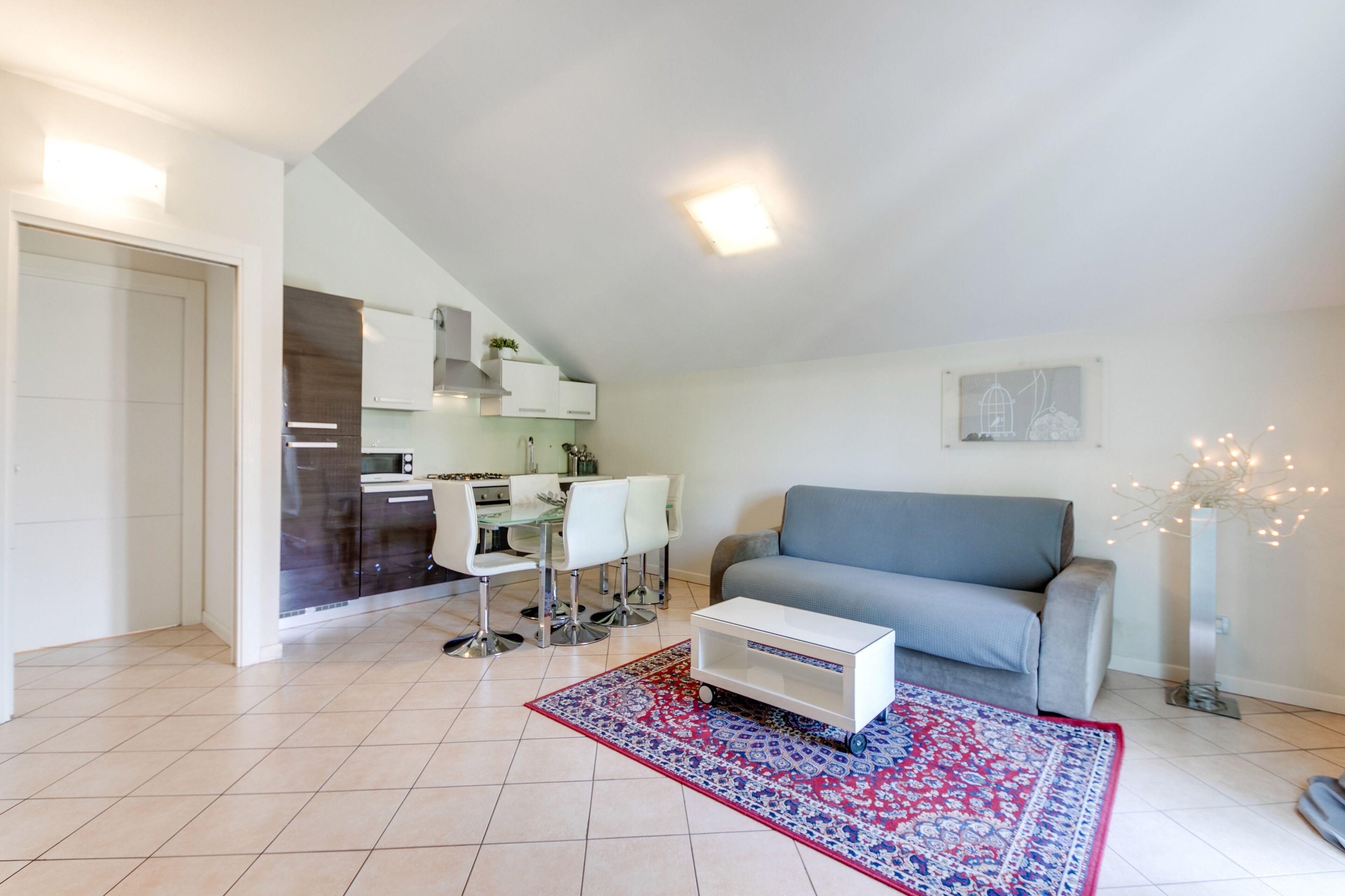 A modern living room with a grey sofa, a small white table, and a patterned rug. There is a kitchen area with white cabinets and a dining table with chairs.