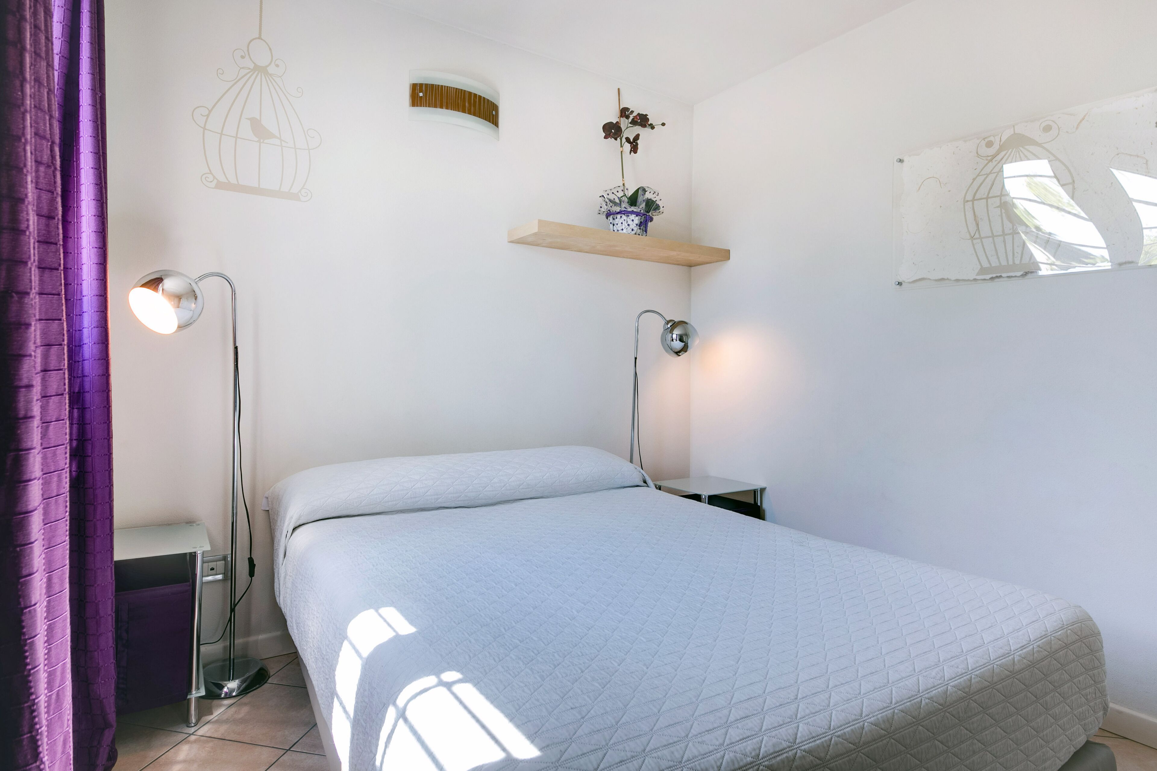 Ein Schlafzimmer mit einem Bett, zwei Nachttischen, einer Stehlampe, einem an der Wand befestigten Regal mit einer Pflanze und einem Fenster mit Vogelkäfig-Design.