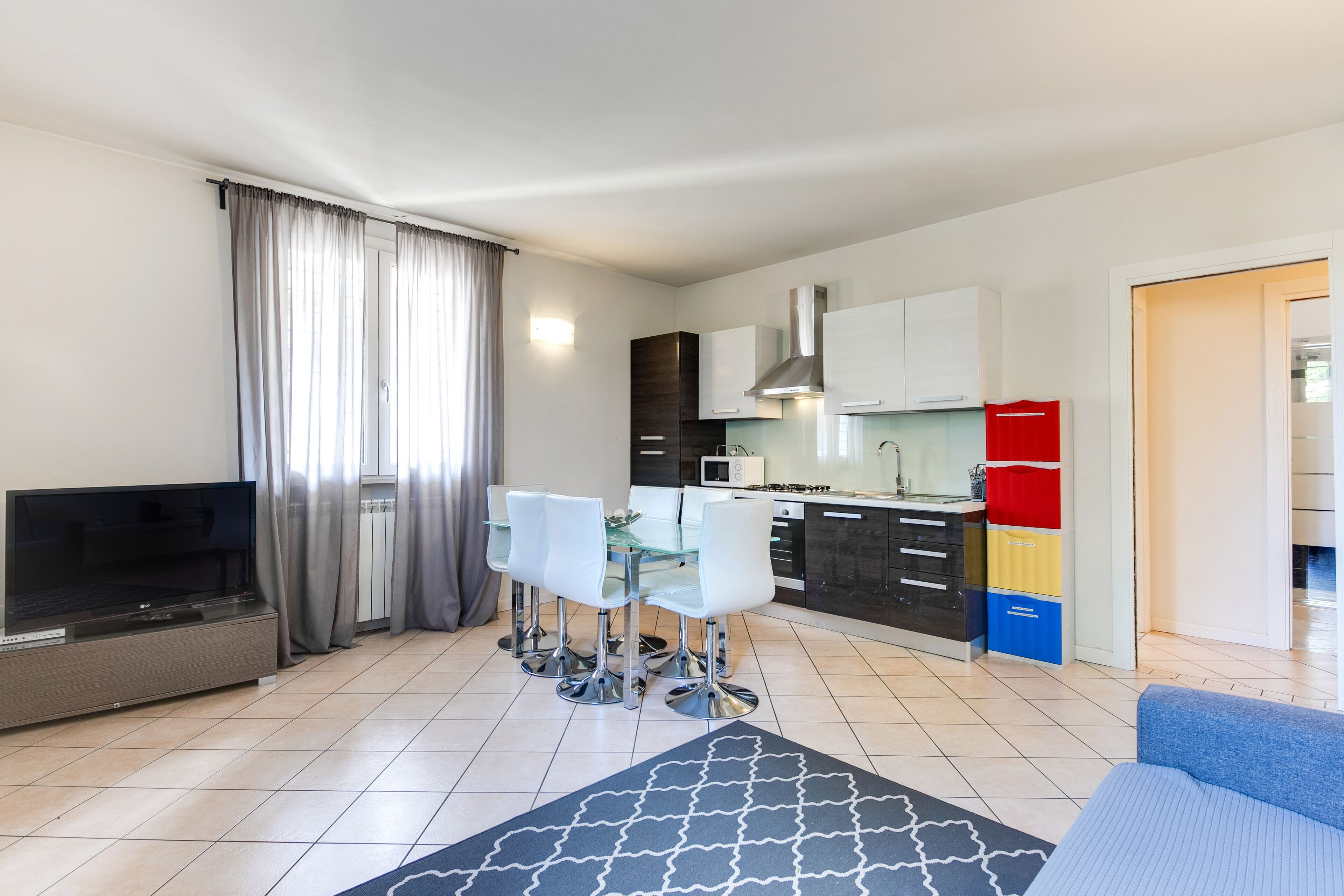 A modern living room with a flat-screen TV, a glass dining table with four chairs, a sofa, and a kitchen area with cabinets and appliances.