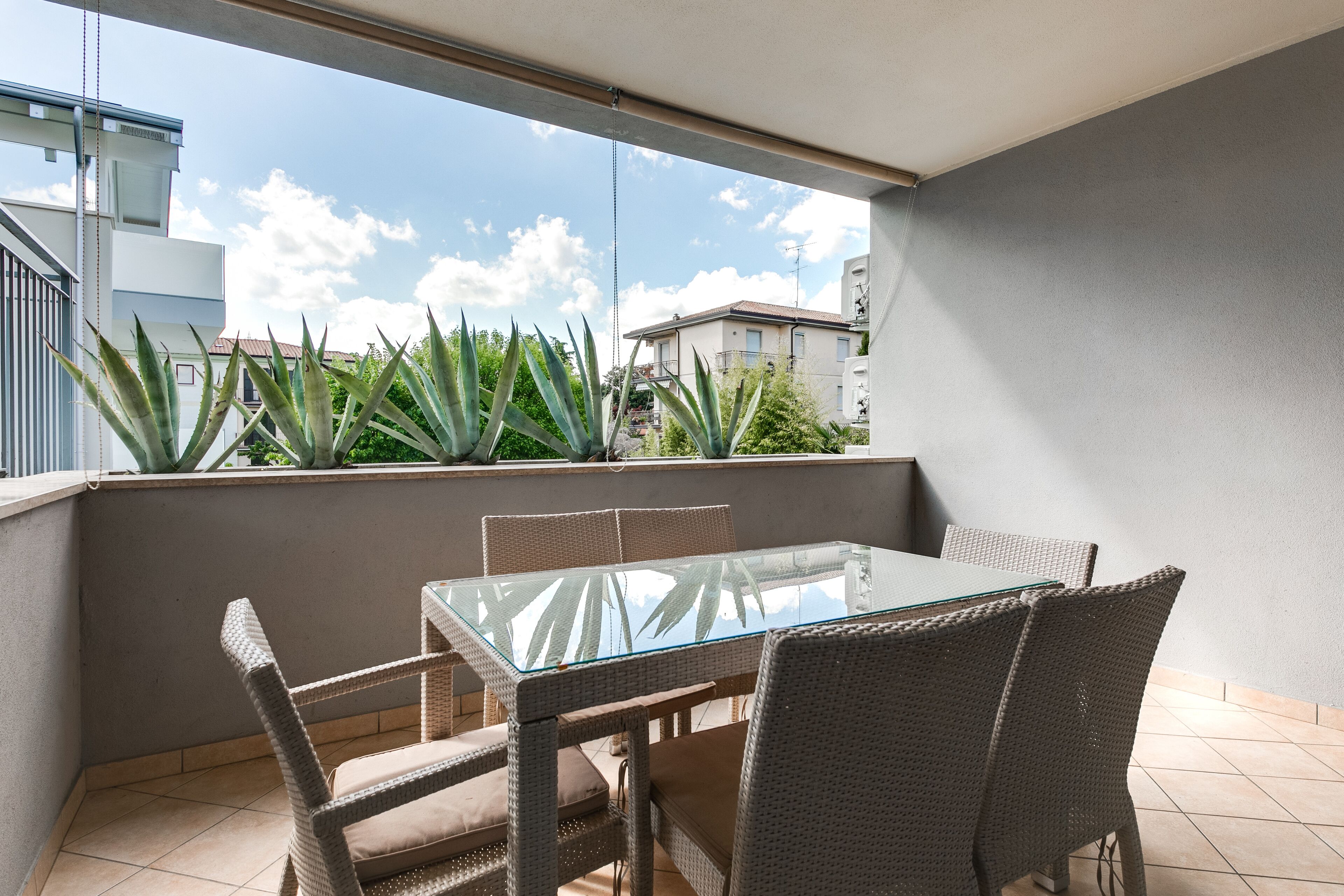 A balcony with a glass table and wicker chairs, overlooking a residential area with buildings and greenery.