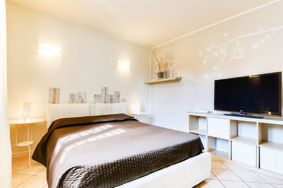 A modern bedroom with a large bed, a flat-screen TV, and a wall-mounted shelf with decorative items.