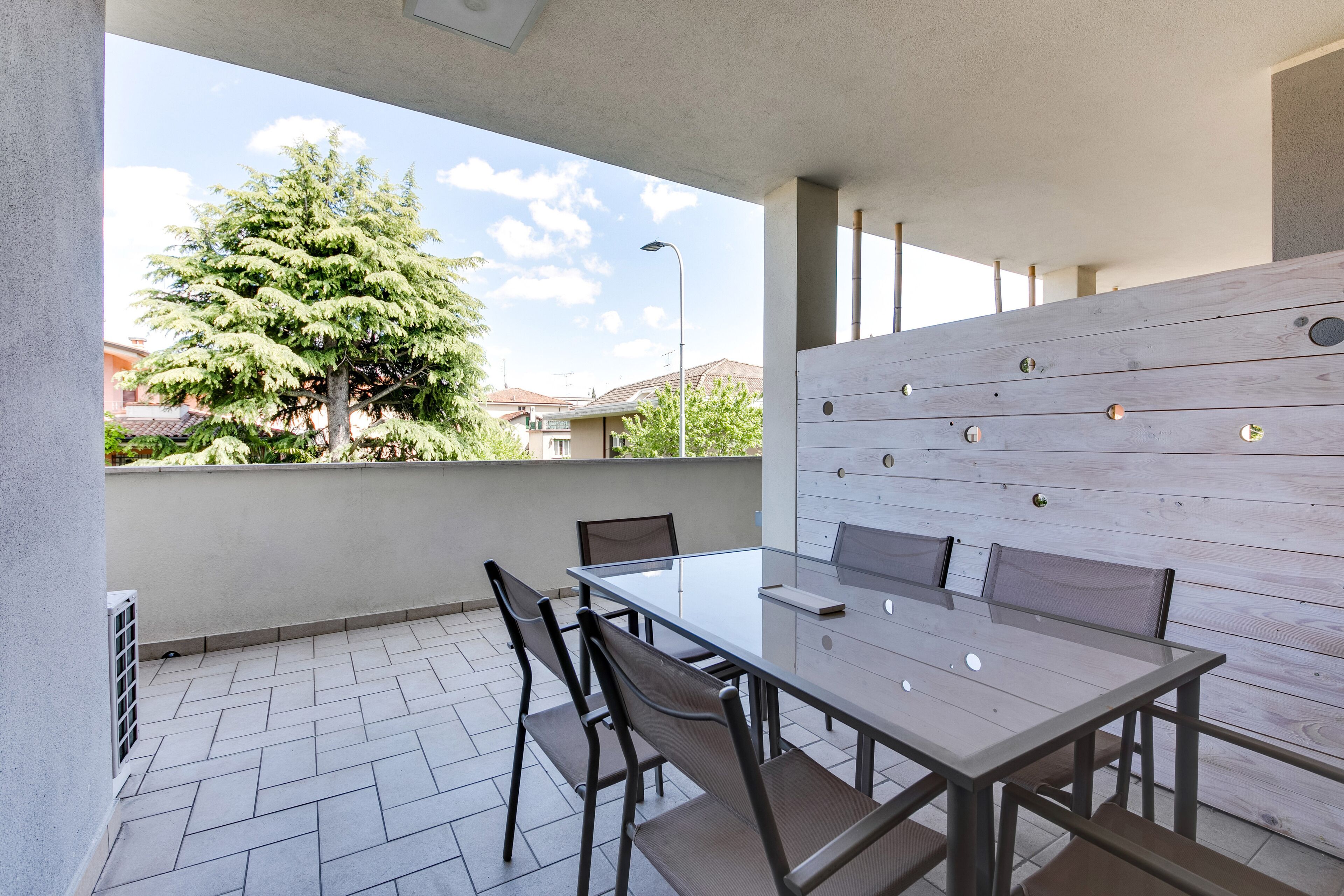 Une terrasse avec une table et des chaises en verre, un mur en bois orné d’ouvertures circulaires, et une vue sur un arbre et des bâtiments en arrière-plan.