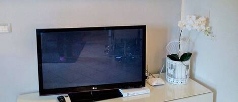 Un televisor de pantalla plana sobre un mueble blanco, con un control remoto y un jarrón decorativo con flores.