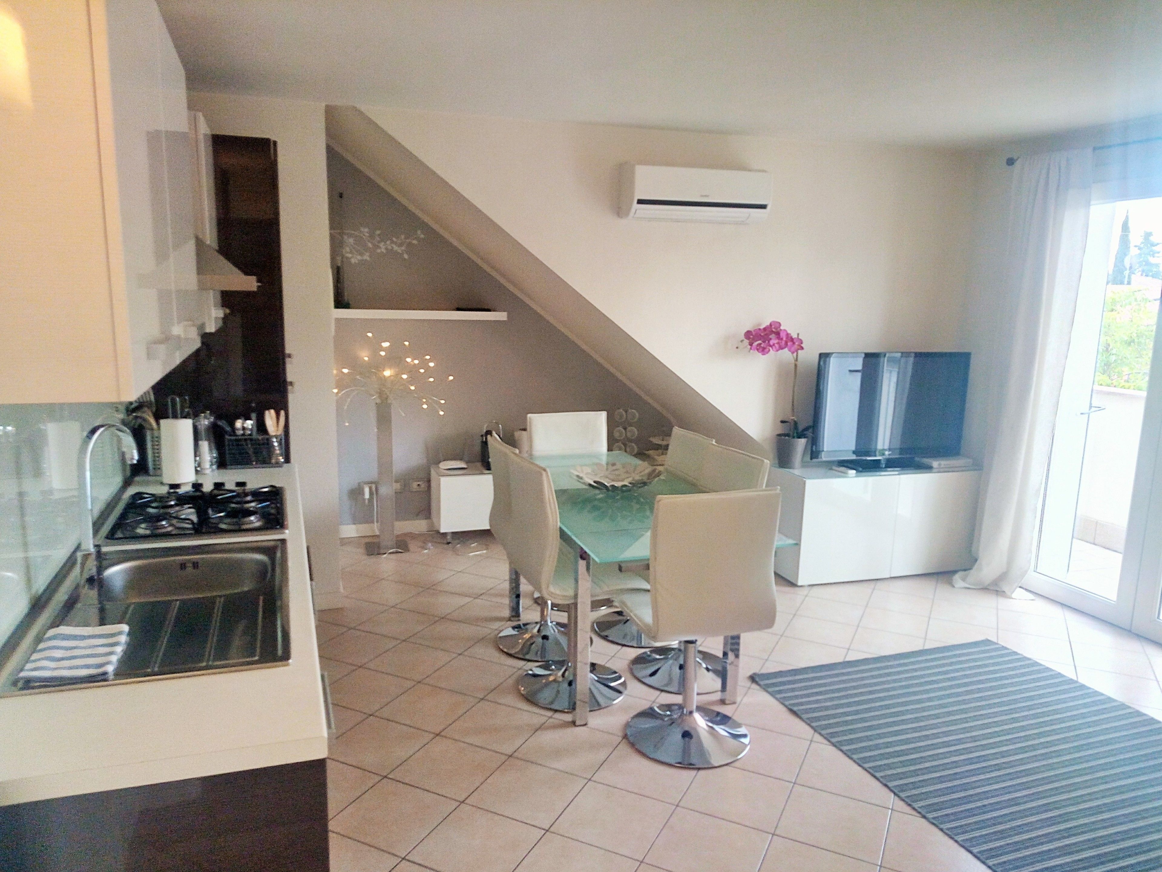 A modern kitchen with a gas stove, a sink, and a dining area with a glass table and chairs. There is a staircase leading to a lofted area with a TV and a potted plant.