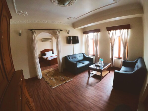 A hotel room with a wooden floor, a black leather sofa, a coffee table with water bottles, a TV mounted on the wall, and a bed with a wooden headboard. - Hotel Bodhiz (Kathmandu)