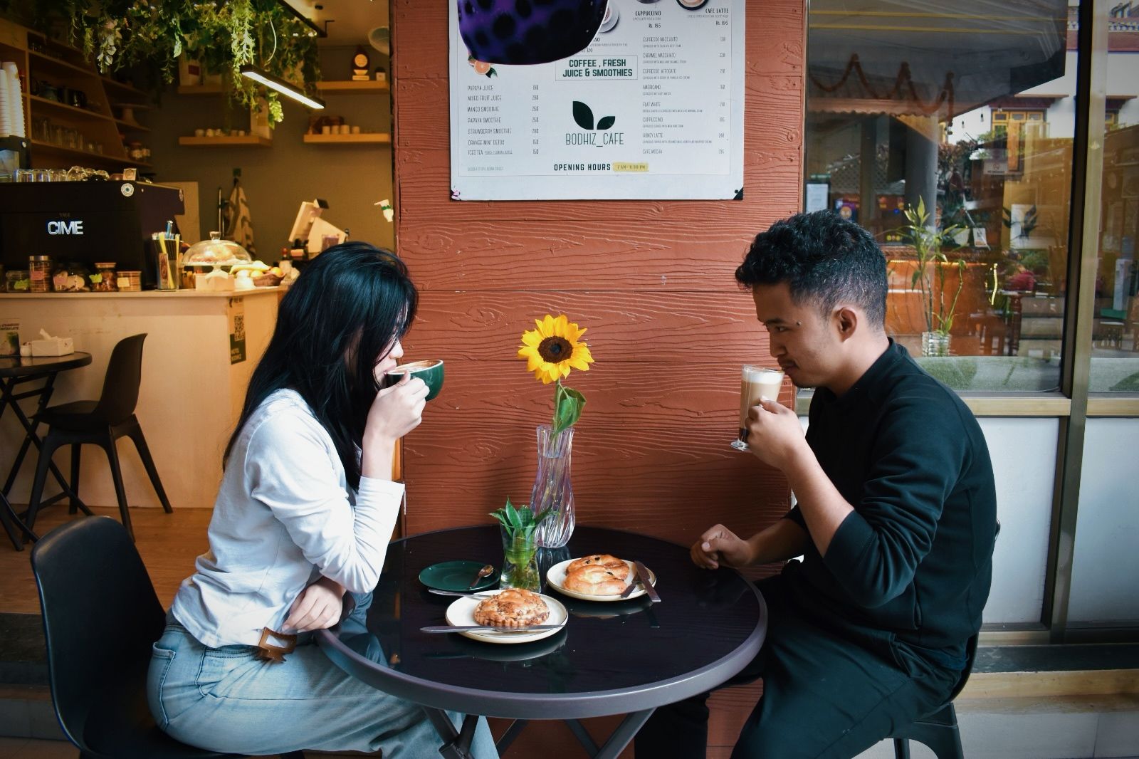 Dos personas sentadas en una mesa redonda con un tablero de menú encima, una máquina de café al fondo y una ventana que ofrece vistas a la calle.