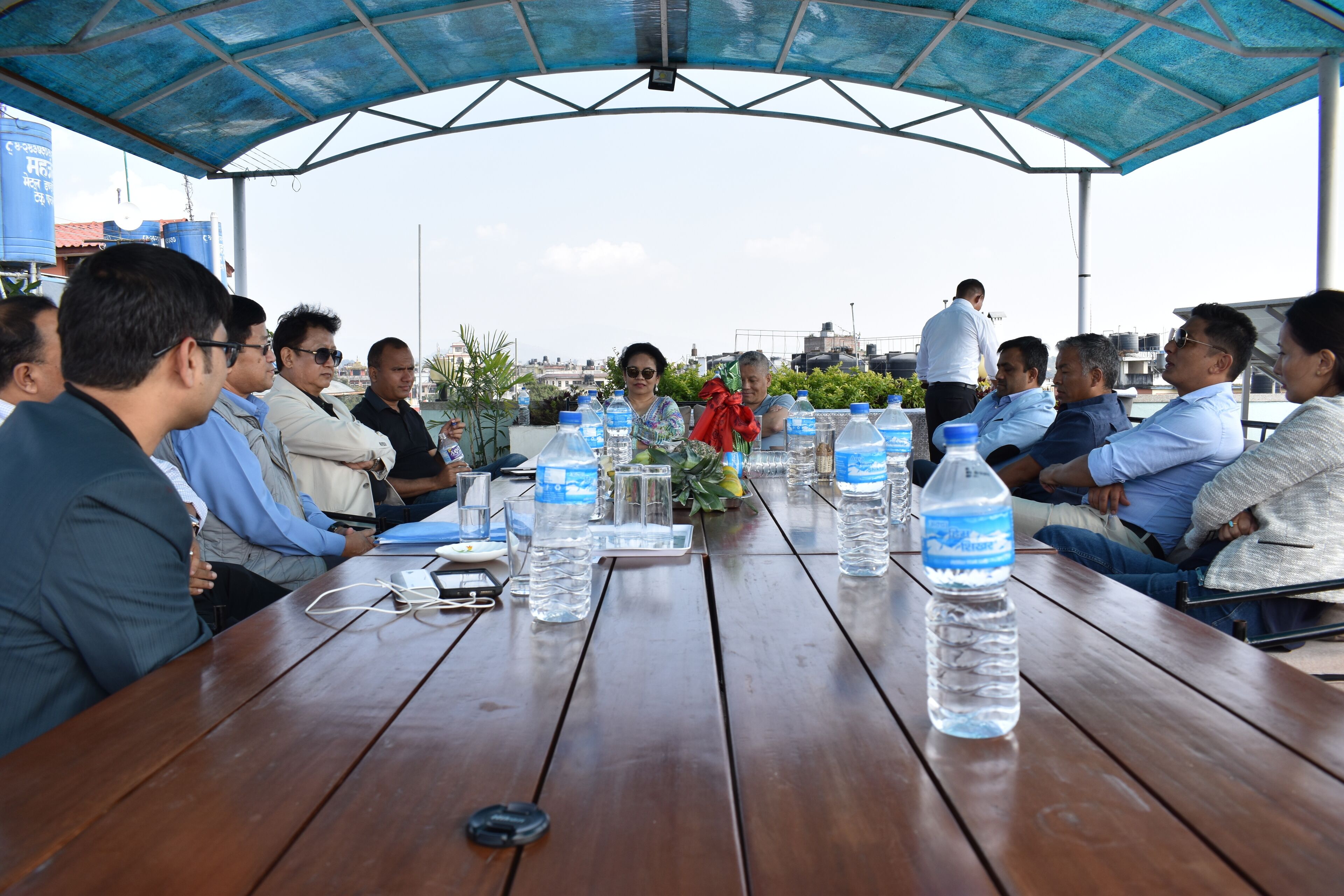 A group of people seated around a wooden table with water bottles and documents, under a blue canopy.