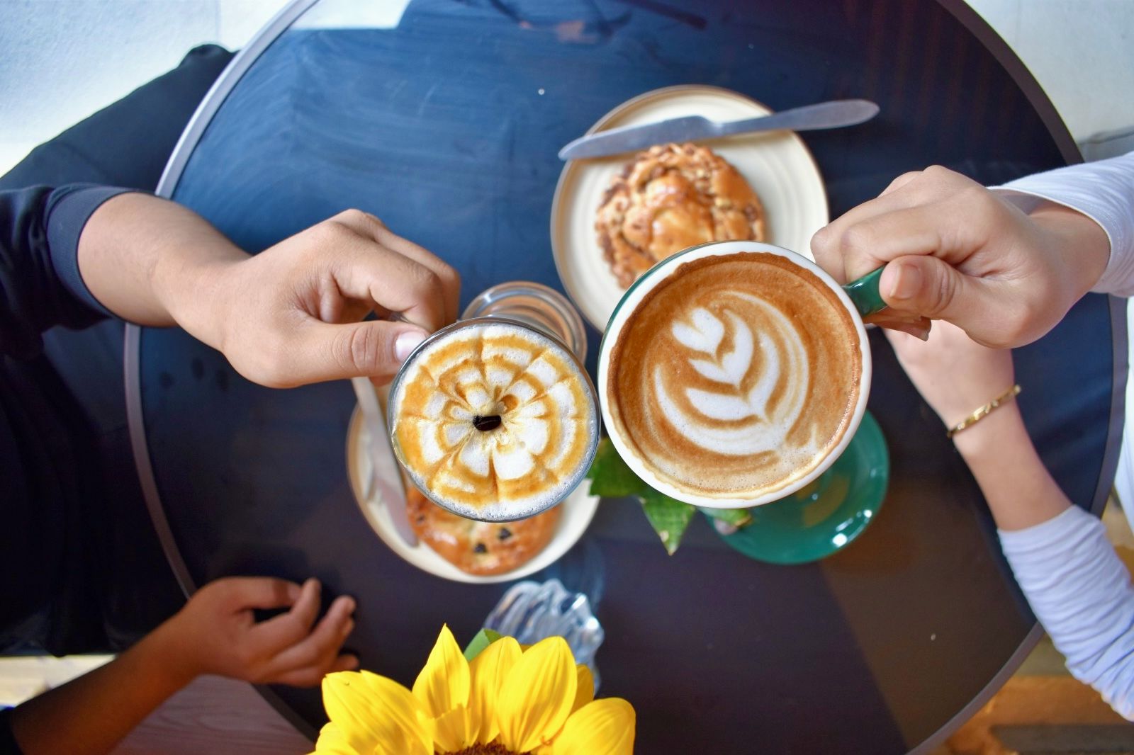 Una mesa redonda con dos tazas de café y un pastel, con manos sosteniendo las tazas.