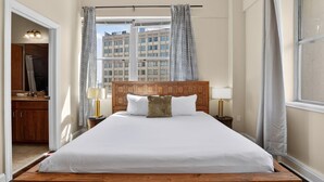 Un dormitorio con cabecera de madera, una cama grande, dos mesitas de noche con lámparas, un ventanal con cortinas estampadas y vistas a edificios.