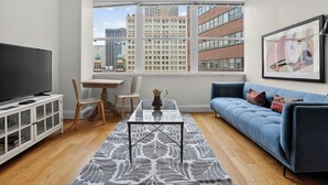 Un moderno salón con televisor, un sofá azul, una mesa de centro y vistas de edificios de la ciudad a través de amplios ventanales.