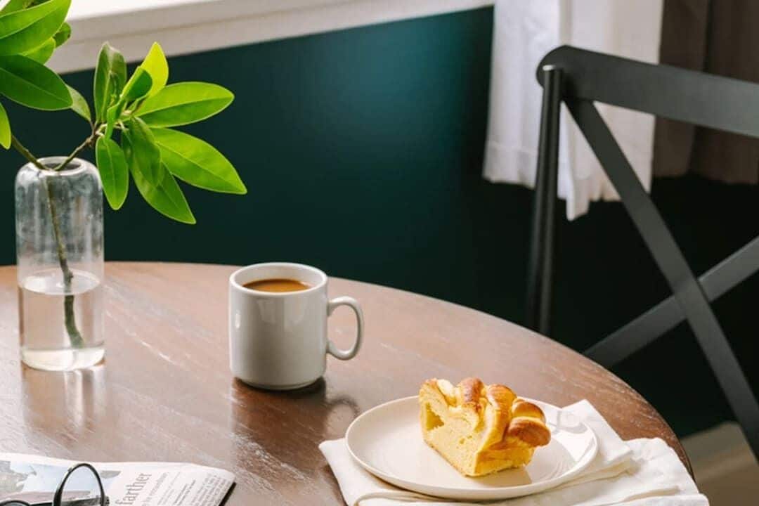 A round wooden table with a white mug, a plate with a croissant, a glass of water, and a newspaper.