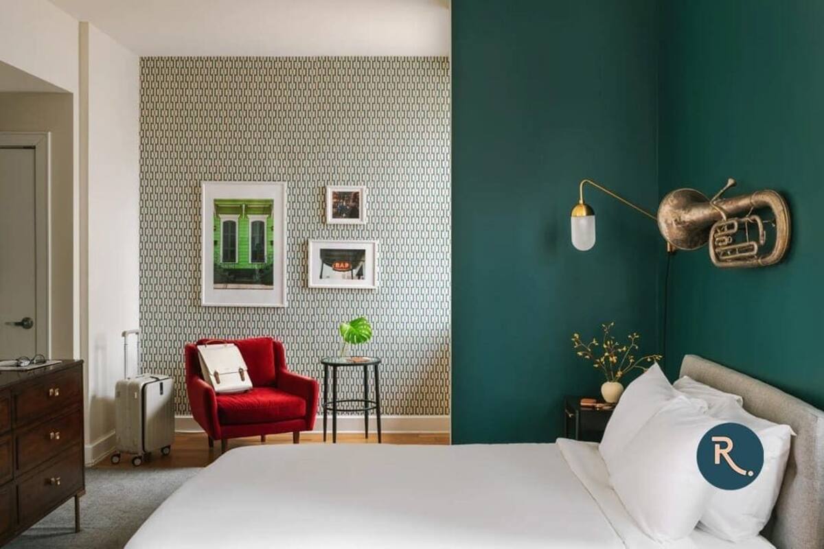 Ein Hotelzimmer mit einem Bett, einem roten Sessel, einem kleinen Tisch mit Pflanze, einem Koffer und einer Wand mit gemustertem Tapeten und gerahmten Bildern.