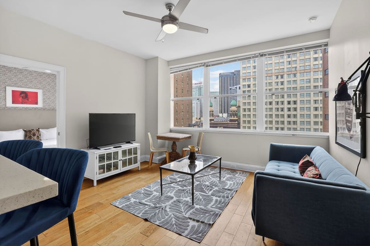 Ein modernes Wohnzimmer mit Fernseher, Sofa, Sesseln, einem Esstisch und einem großen Fenster mit Blick auf die Stadt.