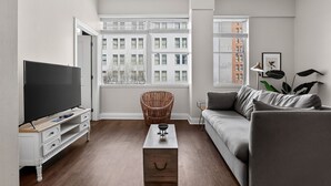 Una sala de estar moderna con una televisión de pantalla plana, un sillón de mimbre, un sofá gris y vistas de edificios a través de amplias ventanas.