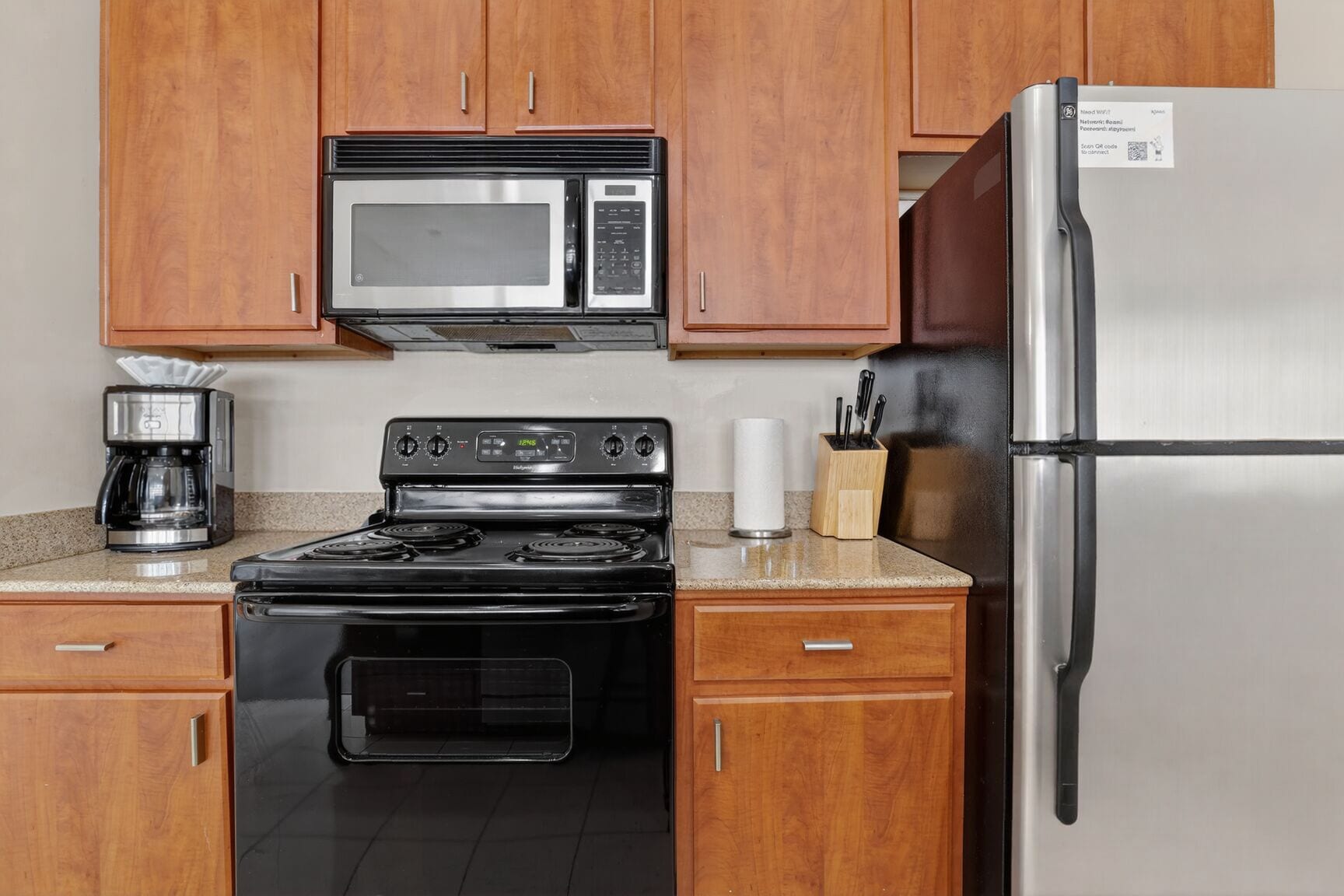A kitchen with wooden cabinets, a stainless steel refrigerator, a microwave, a stove, and a coffee maker.